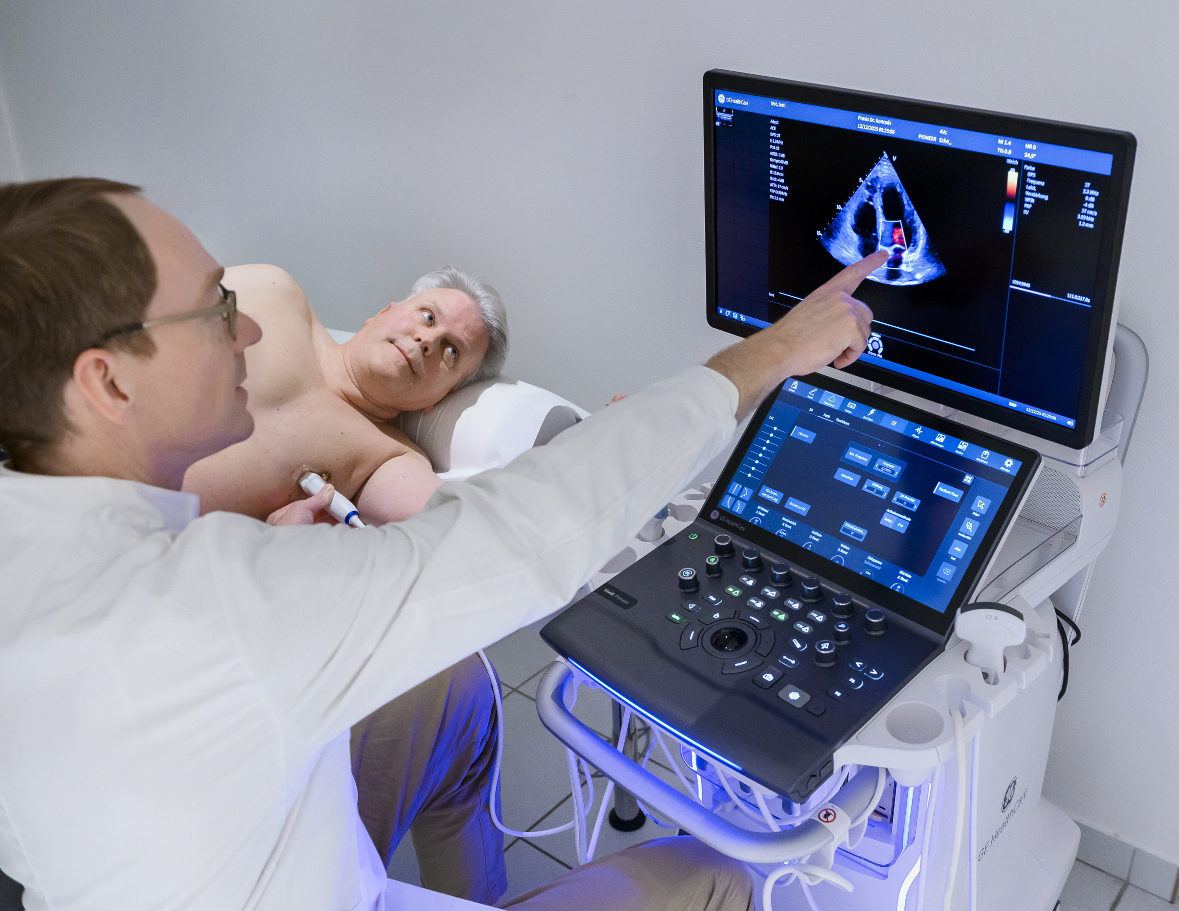 Ein Arzt deutet auf den Monitor eines Ultraschallgeräts. Der Patient blickt ebenfalls auf den Monitor.